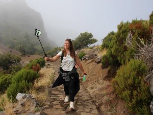 Vrouw wandelt over een bergpad met groene struiken en mistige bergen op de achtergrond terwijl ze een selfie maakt met een selfiestick.