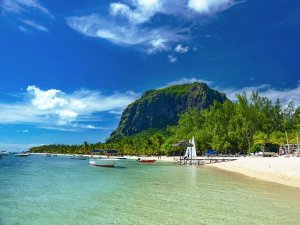 Witte zandstrand met helderblauw water en kleine bootjes, omringd door palmbomen en met de imposante Le Morne Brabant berg op de achtergrond in Mauritius.