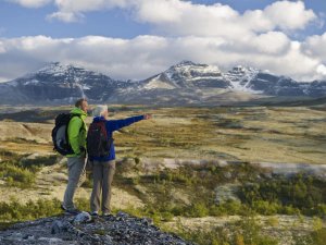 Two hikers on a rocky outcrop overlook a vast mountain landscape, with one pointing into the distance.