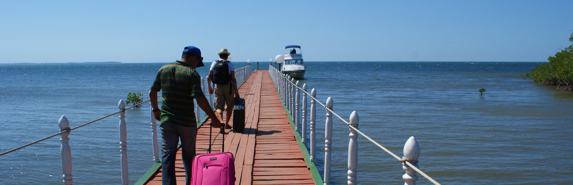 Twee mensen met bagage lopen over een houten pier richting een boot op een zonnige dag in Cuba