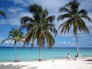 Persoon zit op een wit zandstrand met palmbomen en een turquoise zee onder een blauwe lucht in Cuba