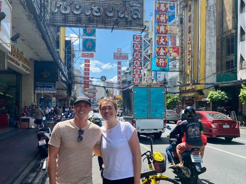 Een stel staat midden in het bruisende Chinatown van Bangkok, omringd door kleurrijke uithangborden in Chinese karakters, brommers, taxi’s en straatverkopers.