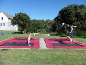 Kind staat op een trampoline terwijl een man springt en een bal kopt op een tweede trampoline in een zonnige tuin in Zuid-Afrika