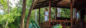 Man in badjas met een kopje koffie in zijn hand staand op een balkon van zijn houten jungle bungalow uitkijkend over de jungle.
