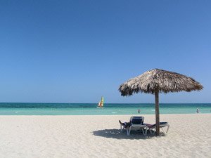 Wit zandstrand met turquoise water, rieten parasol boven ligstoelen en een zeilboot in de verte bij Varadero strand in Cuba