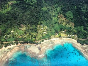 Aerial view of a tropical village on a lush green coast with a sandy beach and turquoise ocean.