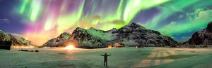 Felgroen en roze noorderlicht verlicht een besneeuwd berglandschap terwijl een persoon met gespreide armen op het ijs staat in IJsland