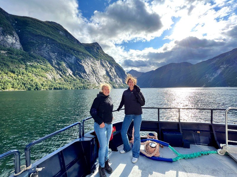 Two women stand on a boat deck in a scenic fjord with mountains and sunlight reflecting on the water.