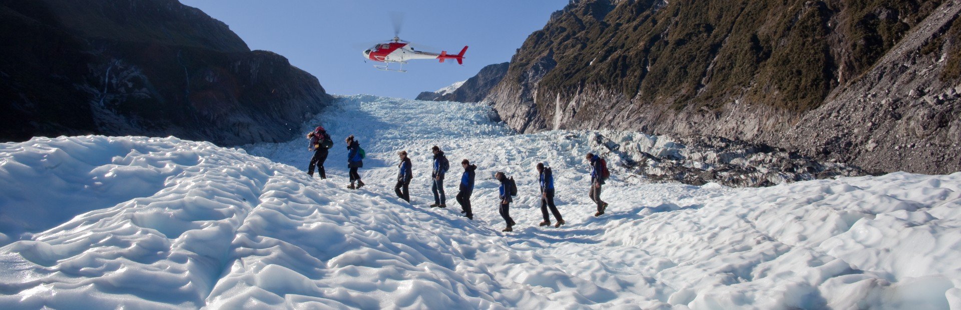 Wandelaars lopen over een gletsjer met een helikopter erboven en bergen op de achtergrond in Nieuw-Zeeland