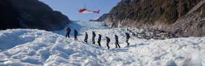 Wandelaars lopen over een gletsjer met een helikopter erboven en bergen op de achtergrond in Nieuw-Zeeland