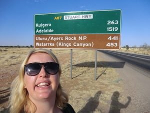 Lachende blonde vrouw met zonnebril staat voor een groot groen bruin afstandsbord met bestemmingen zoals Uluru Ayers Rock NP en Adelaide langs de Stuart Highway in de Australische outback