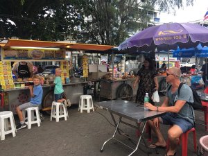 Straatscène bij een Thaise food market, met kinderen op krukjes bij een eetkraam en volwassenen aan een tafel onder een paarse parasol.