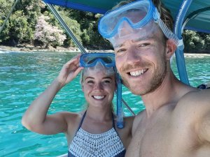 Twee reizigers met snorkelmaskers poseren lachend op een boot boven helderblauw tropisch water.