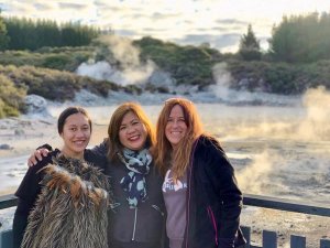 Drie lachende vrouwen, waarvan één een traditioneel veren kledingstuk draagt, poseren voor een stomend geothermisch landschap in een vulkanisch natuurgebied