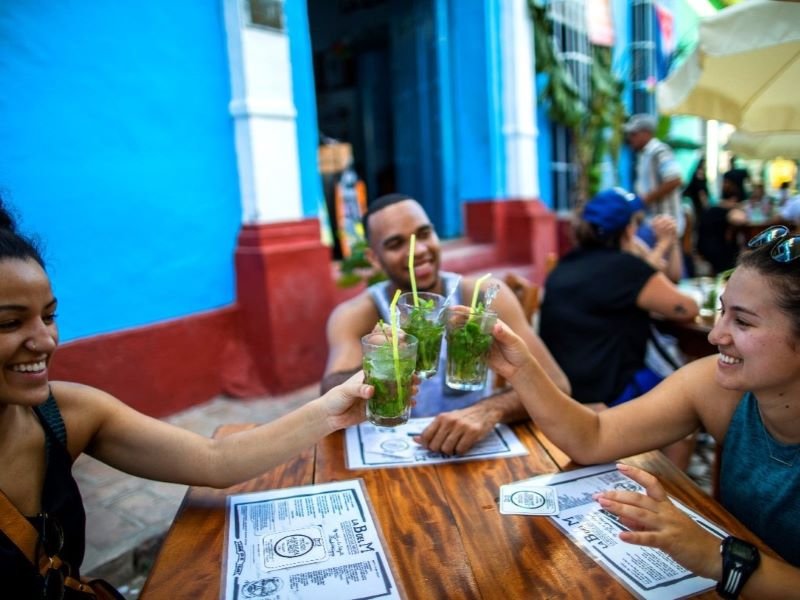 Drie glimlachende mensen proosten met glazen muntdrankjes op een buitencafé in Cuba
