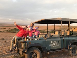 Gezin van vier poseert vrolijk op een safariwagen van Garden Route Game Lodge in een droog, open landschap in Zuid-Afrika