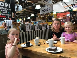 Vrouw en twee jonge meisjes genieten van drankjes en desserts aan een houten tafel in een druk café in Zuid-Afrika