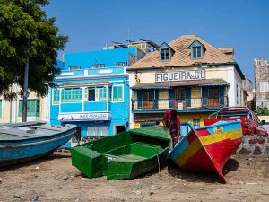 Persoon werkt op een groene boot op een zandstrand tussen andere kleurrijke boten met felblauwe en gele gebouwen op de achtergrond onder een heldere lucht aan de kust