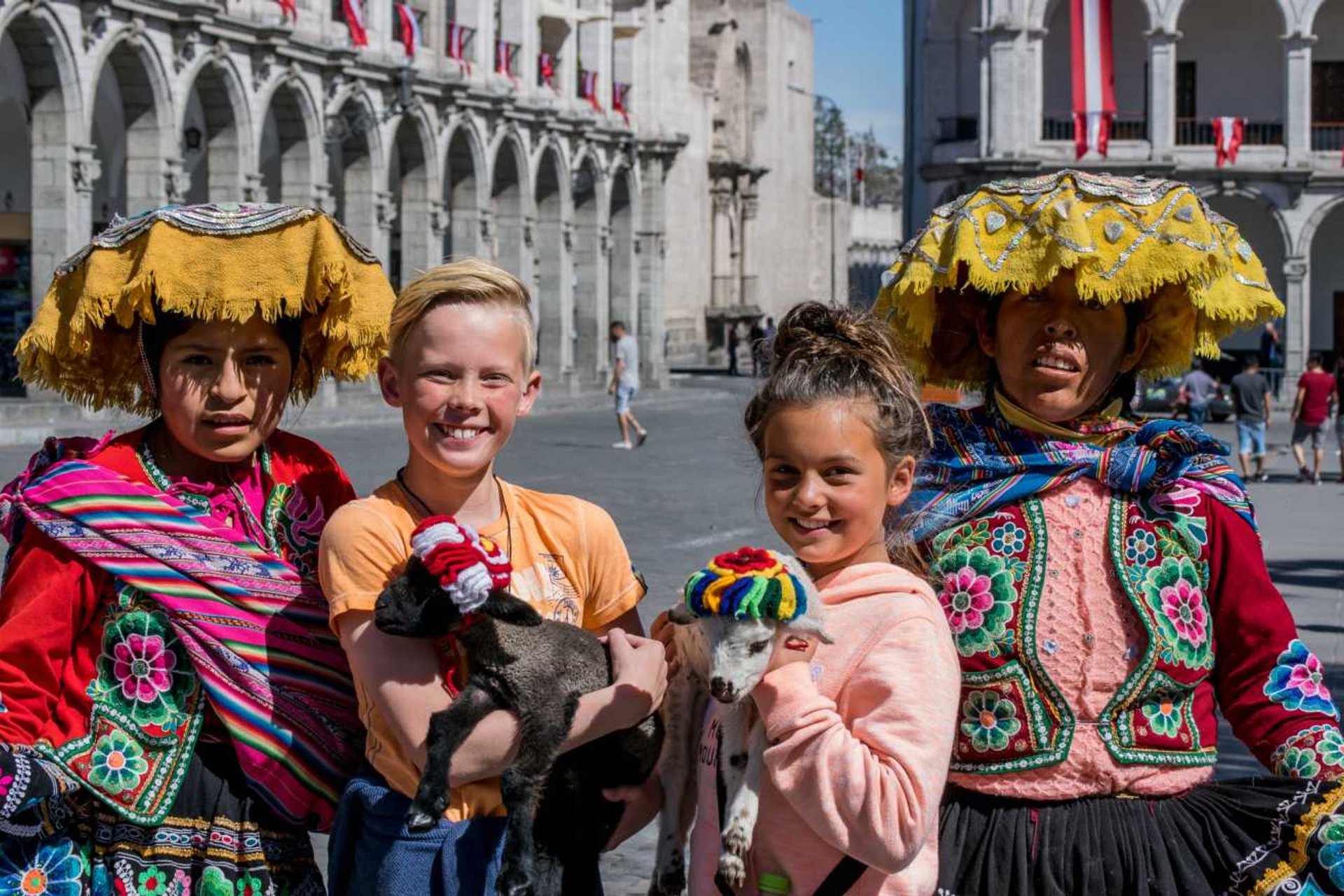Twee kinderen poseren met twee Peruaanse vrouwen in kleurrijke traditionele kleding en lammetjes op een plein.