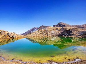 Kleurrijk geothermisch meer weerspiegelt rotsachtige stomende vulkanische bergen onder een heldere blauwe lucht in een vulkanisch natuurgebied
