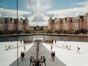 A modern glass facade reflects older buildings, a canal, and a white plaza bustling with people, creating a mirrored scene.