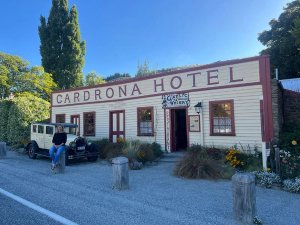 Vrouw zit op een houten paal voor het historische Cardrona Hotel met een vintage auto geparkeerd in een landelijk gebied in Nieuw-Zeeland
