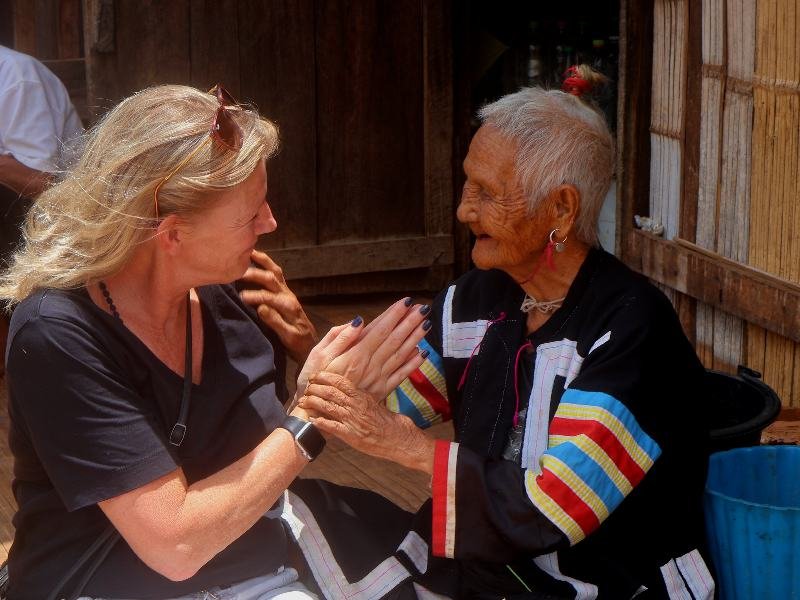 Een blonde reiziger zit op de grond tegenover een oudere vrouw in traditionele kleurrijke kleding. Ze houden elkaars handen vast en lachen hartelijk naar elkaar. Op de achtergrond is een houten hut te zien en zit een man op blote voeten.