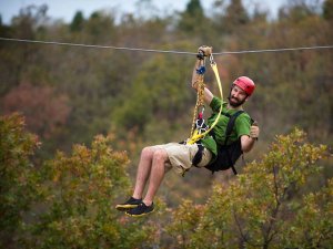 Man met baard en rode helm in Albanië die glimlacht en een duim omhoog geeft tijdens het ziplinen tussen de bomen