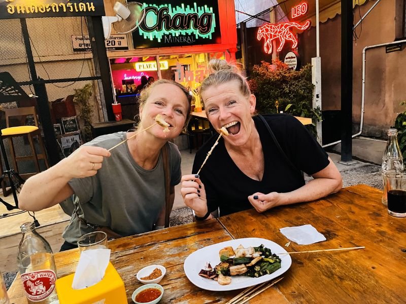 Twee vrouwen zitten lachend aan een houten tafel op een avondmarkt en eten spiesjes. Op tafel staan dipsausjes en een bord met streetfood. Op de achtergrond kleurrijke neonreclames.