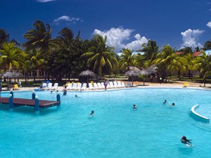 Helderblauw zwembad bij een tropisch resort met zwemmende mensen, palmbomen en rieten hutten onder een heldere lucht in Cuba