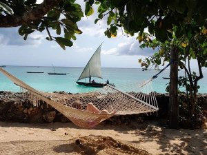 Persoon ligt ontspannen in een hangmat op een zandstrand met uitzicht op turquoise water en een traditionele zeilboot aan de kust