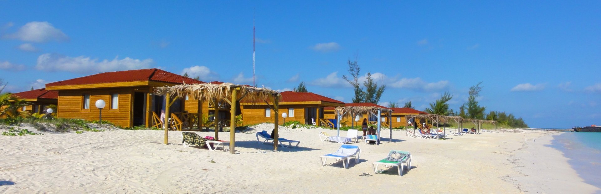 Panorama van houten bungalows met rode daken op een wit zandstrand in Cayo Levisa, Cuba met rieten parasols, ligbedden en turquoise water onder een blauwe lucht