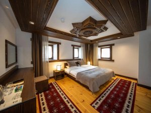 Slaapkamer in Kosovo met tweepersoonsbed, lichte houten vloer, twee rood gedessineerde tapijten, witte muren en donker houten plafond met stervormig ornament