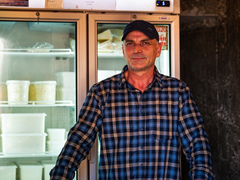 Man in Albanië met blauw geruit overhemd en pet staat voor koelkasten met zuivelproducten