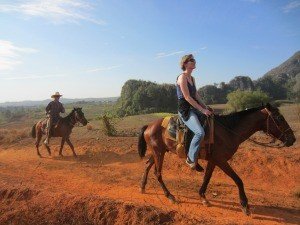 Twee mensen rijden te paard over een rood zandpad met heuvels in een zonnig landschap in Cuba