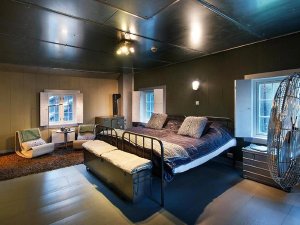 A bedroom with a dark blue bed, large metal fan, and two modern chairs. The room has dark green and light wood walls, a metallic ceiling, and two open-shutter windows.