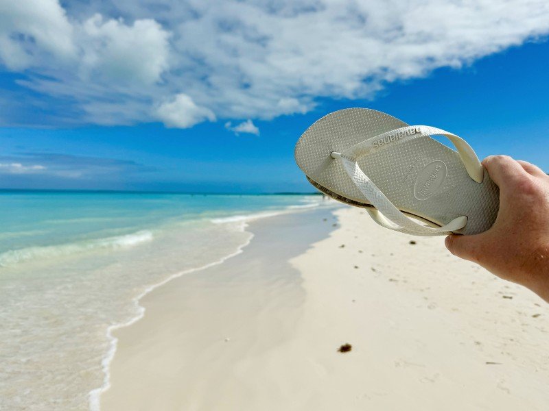 Een hand houdt een witte slipper vast tegen de achtergrond van een wit zandstrand en een turquoise zee onder een blauwe lucht.