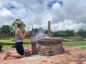 Een vrouw knielt op een stenen plateau bij een roodbruine bakstenen offerplaats in het tempelcomplex van Sukhothai, Thailand. Ze houdt wierookstokjes vast in een gebedsgebaar. Op het altaar liggen kleurrijke offers, waaronder gele en roze bloemen, bananen en ander fruit. Wierookrook kringelt omhoog. Op de achtergrond zijn eeuwenoude tempelruïnes en stoepa’s te zien, omringd door helder groen gras en bomen onder een lichtbewolkte hemel.