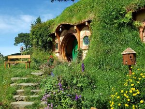 Hobbitwoning ingebouwd in een grasheuvel met een ronde groene deur, stenen trap, houten bank en kleurrijke bloemen in een filmdecor in Nieuw-Zeeland