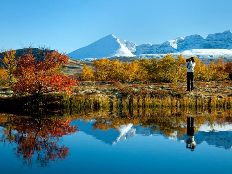 A person photographs a scenic autumn landscape with colorful trees and snow-capped mountains reflecting in a lake.