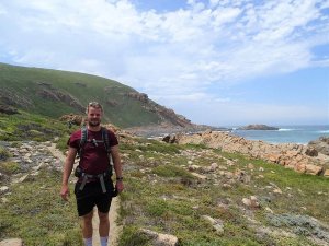 Man met rugzak staat op een wandelpad langs de kust met groene heuvels en uitzicht op een rotsachtige oceaan in Zuid-Afrika