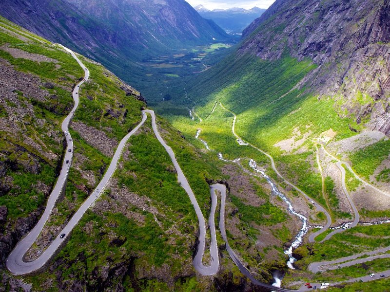 A winding mountain road with hairpin turns descends a steep, green valley with a river.