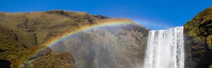 Regenboog buigt over een krachtige waterval die langs groene kliffen naar beneden stroomt onder een blauwe lucht in IJsland