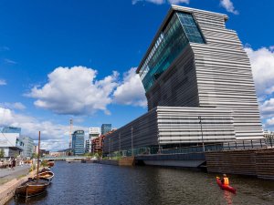 A modern, grey, ribbed building stands beside a city canal with moored boats and a person in a red kayak, under a blue sky with white clouds.