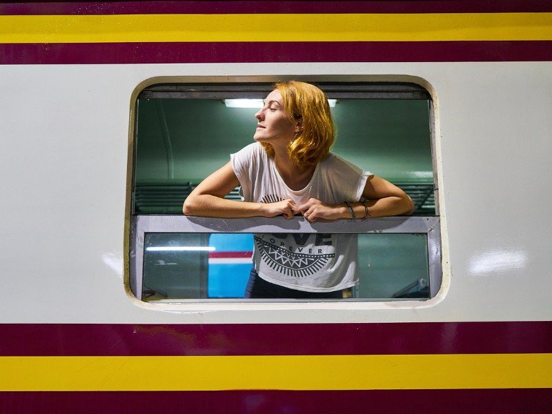 Vrouw met halflang rossig haar leunt met beide armen op het raamkozijn van een wit-rode trein met gele streep.
