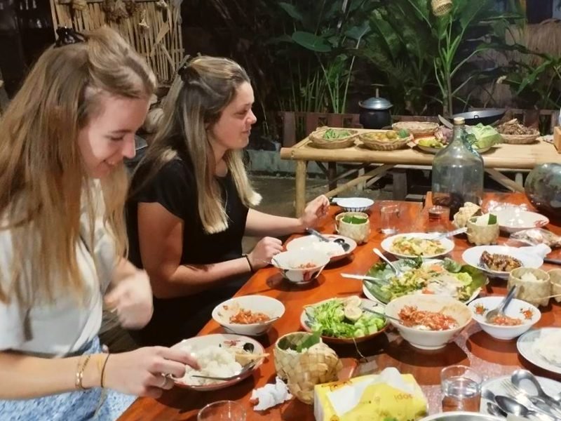 Twee vrouwen zitten aan een houten tafel. Op tafel staan allerlei schalen en borden met lokale gerechten. Op de achtergrond staan manden met verse ingrediënten op een bamboetafel. De setting is buiten, omringd door grote groene planten.