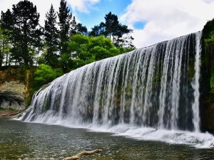 Brede waterval stort in een rivier met groene bomen en rotsachtige oevers onder een blauwe lucht in een natuurgebied