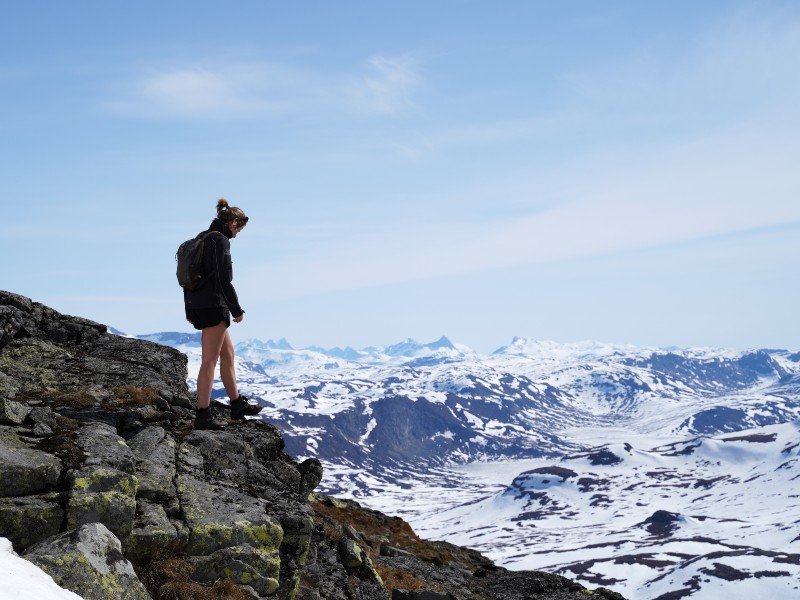 Woman with backpack on a rocky cliff overlooking a snowy mountain range.