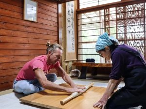 Man en oudere vrouw rollen samen deeg uit op een houten plank tijdens een kookles in Japan.