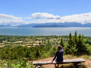 Persoon zit op een bankje met uitzicht over een uitgestrekt meer, groene velden en bergen in de verte bij Ohrid in Noord-Macedonië